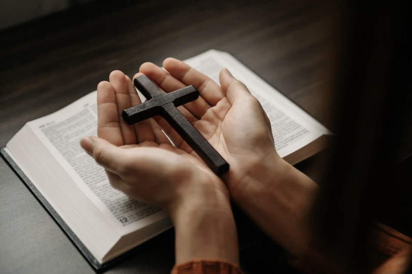 A woman holds a wooden cross in her hands while resting them on an open Bible.