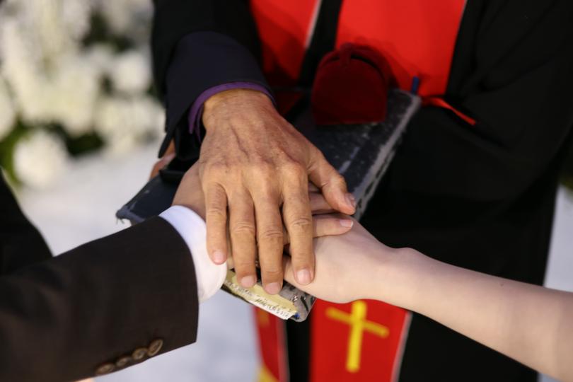 A pastor places his hand over those of a bride and groom during a Christian wedding ceremony on an unknown day.