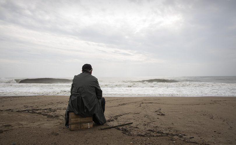 A man sits alone on a small stool by the seaside under a cloudy sky on an unknown day.