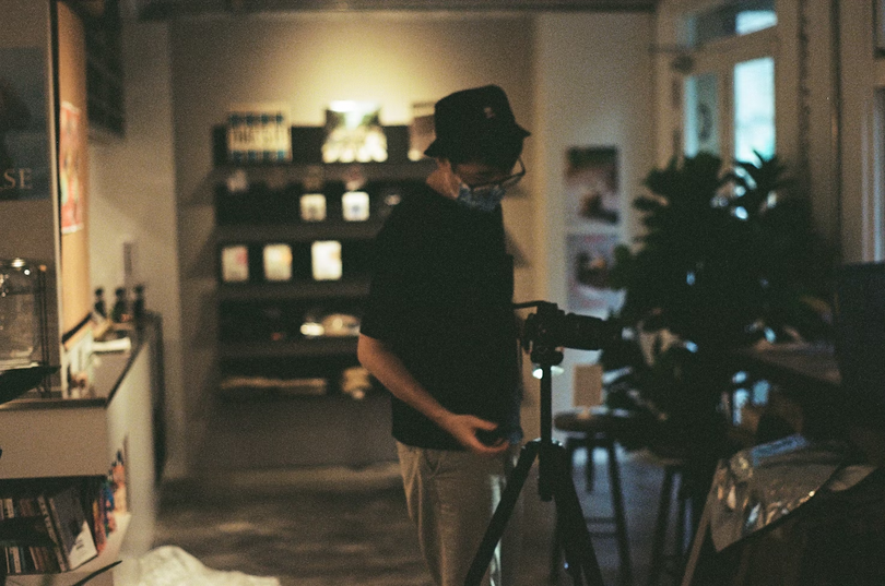 A man in a black shirt and brown pants stands near a black camera tripod on an unknown day.