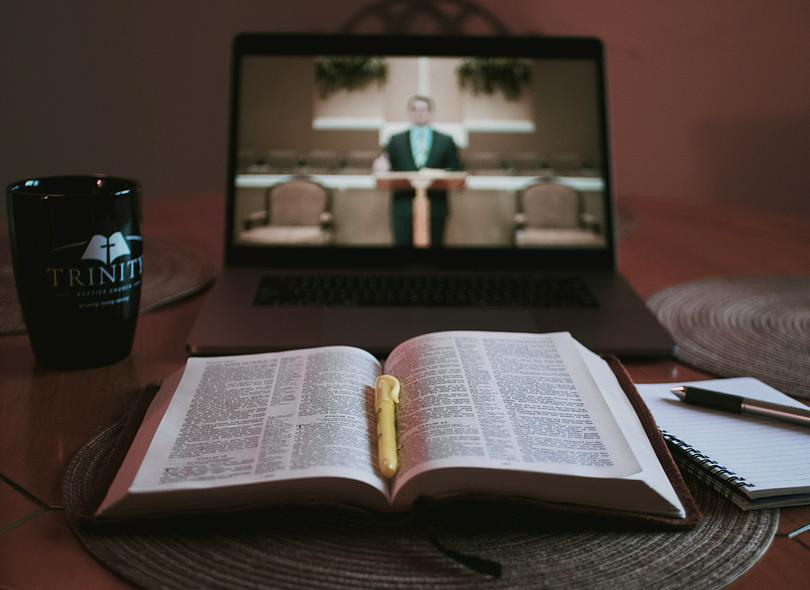 A laptop streaming a virtual sermon sits in front of an open Bible on a wooden surface.