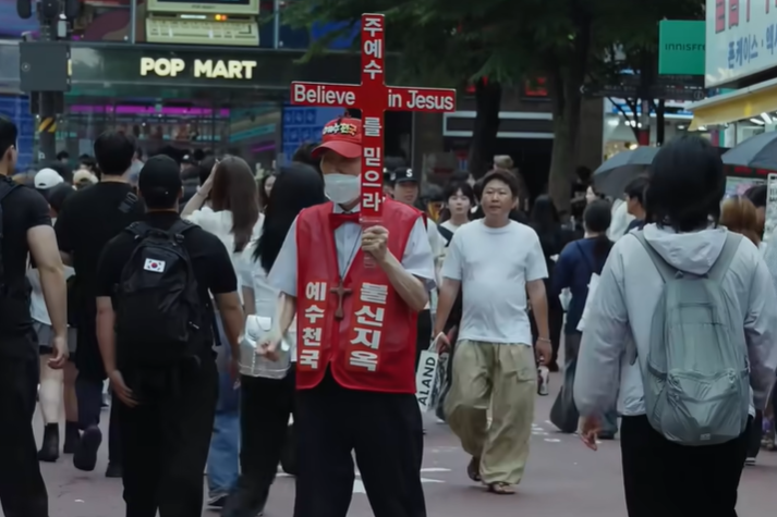 A man wearing a red vest emblazoned with the Korean phrases “Jesus Heaven” and “Not Believe Hell” held up a red cross displaying “Believe in Jesus” in both English and Korean on a crowded street on an unknown day.