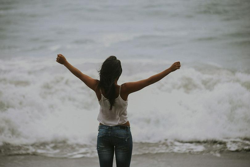 A woman stands by the sea with opening arms.
